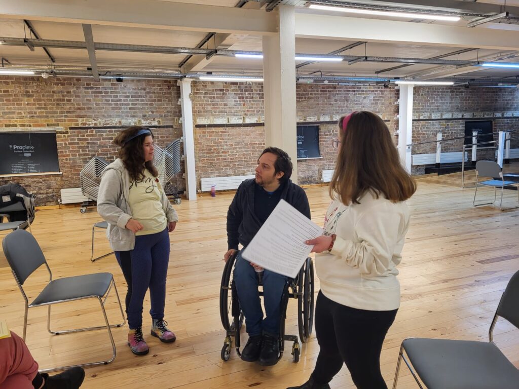 Thom chats to two participates wearing leggings and jumpers. One holds a script. They are surrounded by grey chairs. In the background, an exposed brick wall can be see.